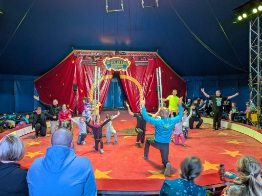 Zirkusaufführung mit Kindern und Artisten in bunten Kostümen auf einer roten Bühne.