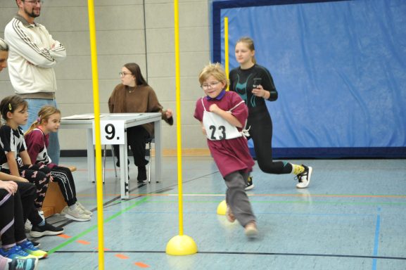 Ein Kind sprintet in einer Sporthalle zu einem Ziel, während ein weiteres Kind folgt.