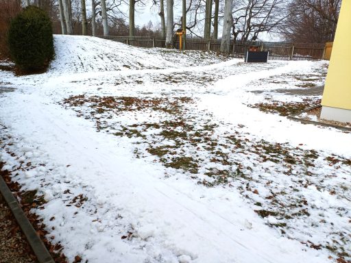 Schneebedeckter Garten mit leicht sichtbaren Grasflächen und Bäumen im Hintergrund.