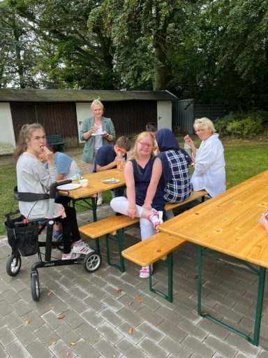 Gruppenszene im Freien, Personen sitzen an Holztischen und unterhalten sich.