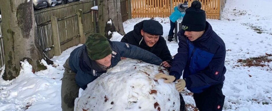 Drei Personen rollen einen großen Schneeball im Schnee, umgeben von verschneiten Landschaft.