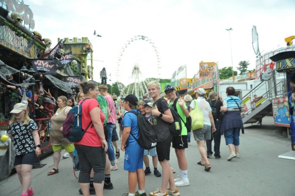 Menschenmenge auf einem Jahrmarkt, mit Ständen und Fahrgeschäften im Hintergrund.