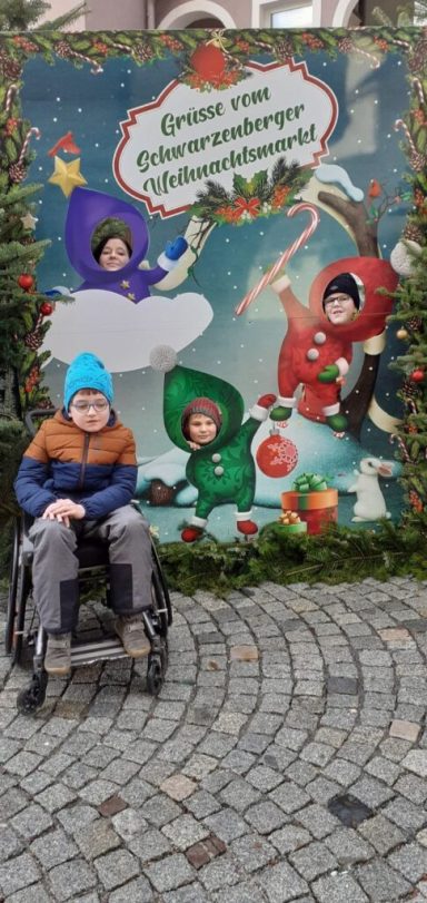 Kind im Rollstuhl vor einem weihnachtlich dekorierten Hintergrund mit vier fröhlichen Figuren.