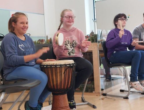 Zwei Mädchen spielen auf einer Djembe, während andere im Hintergrund musizieren.