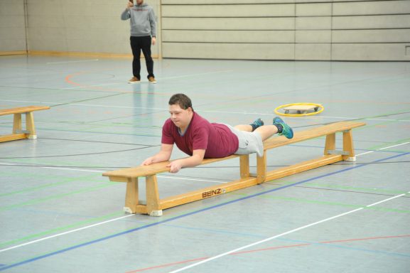 Junge liegt auf einer Bank in einer Sporthalle und übt eine Bewegung.