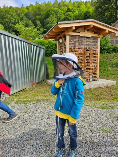 Junge in blauer Jacke und Hut mit Schutznetz steht vor einer Holzstruktur im Freien.