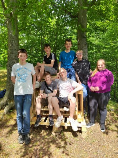 Gruppe von sieben Jugendlichen sitzt auf einer Holzbank im Wald.