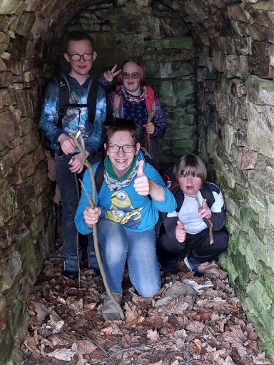Vier Kinder posieren in einem steinernen Tunnel, zwei halten Stöcke und lächeln.