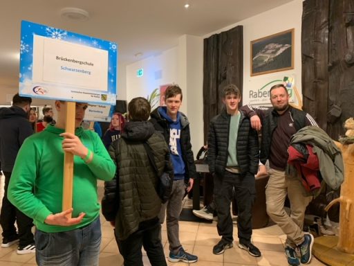 Junge mit Schild steht vor einer Gruppe von Leuten in einem Veranstaltungsraum.