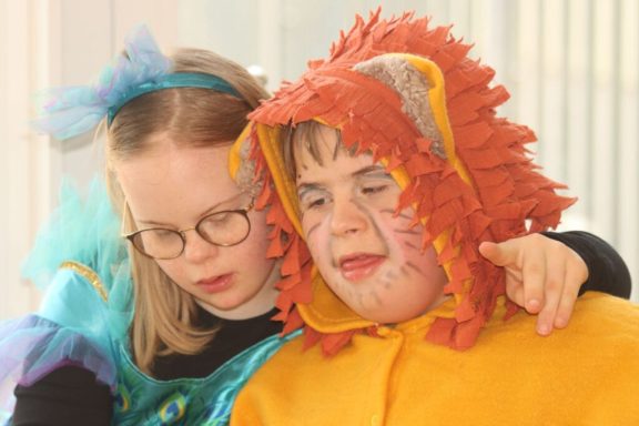 Ein Kind in Löwenkostüm umarmt ein Mädchen in buntem Kostüm, beide lächeln.