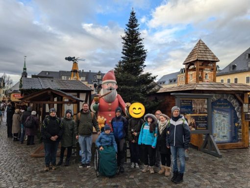 Gruppe von Menschen vor einem Weihnachtsmarkt mit einem großen Weihnachtsmann und Tannenbaum.