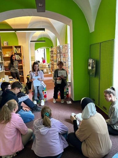 Gruppensitzung in einer Bibliothek mit Menschen auf dem Boden und einer Leserin.