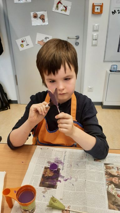 Junge mit braunen Haaren schneidet lila Papier, umgeben von Malutensilien und Zeitungen.