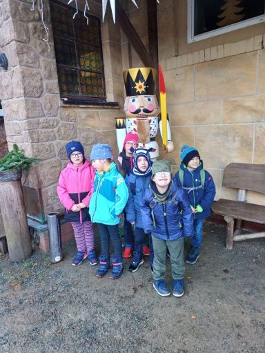Sechs Kinder in farbigen Jacken stehen vor einer hölzernen Weihnachtsfigur.