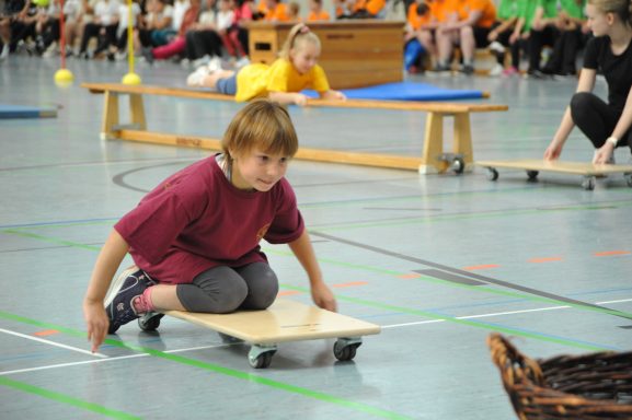 Ein Junge in einem roten Shirt fährt auf einem Rollbrett in einer Sporthalle.