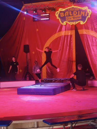 Akrobatin springt von einem Trampolin auf eine Zirkusbühne mit roten Vorhängen.