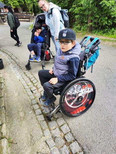 Junge im Rollstuhl mit einer Cap, während er an einem Weg steht, umgeben von anderen Personen.