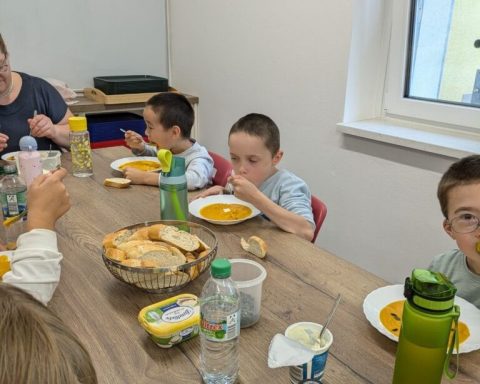 Kinder essen gemeinsam Suppe und Brot an einem Tisch. Eine Lehrerin sitzt dabei.