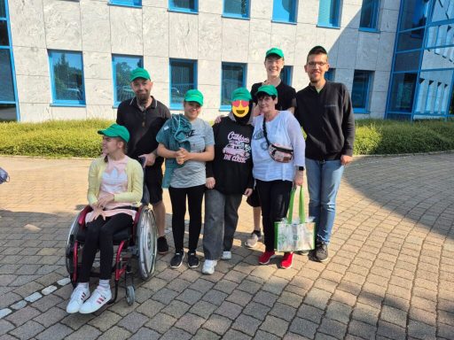 Sechs Personen, darunter ein Mädchen im Rollstuhl, tragen grüne Caps vor einem Gebäude.